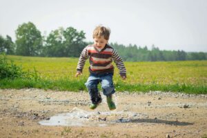 Child wearing gumboots jumping in a muddy puddle, enjoying outdoor play and exploring nature.